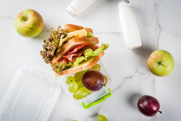 Back to school. A healthy lunch in a box is fresh fruit (apples, plums, grapes), a bottle of yogurt and a sandwich with lettuce, tomatoes, cheese, meat. White marble table. Copy space top view