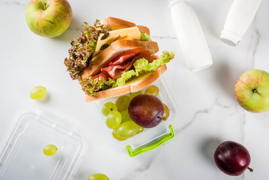 Back to school. A healthy lunch in a box is fresh fruit (apples, plums, grapes), a bottle of yogurt and a sandwich with lettuce, tomatoes, cheese, meat. White marble table. Copy space top view