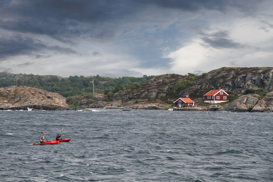Two persons kayaking in the ocean in the coast of Sweden with typical red houses and small rock hills seen in the background with dark and light clouds on the background.