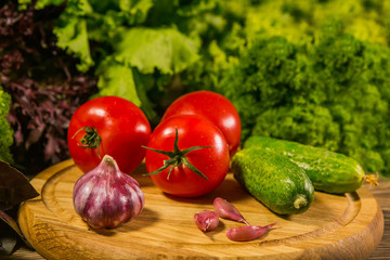 A wooden board with fresh tomatoes, garlic and cucumbers. Green salad in the background.