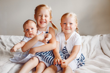 Lifestyle portrait of cute white Caucasian girls sisters holding little baby, sitting on bed indoors. Older siblings with younger brother sister newborn. Family love bonding together concept.