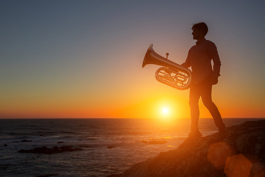 Silhouette of musician with trumpet on rocky sea coast during sunset. Tuba.