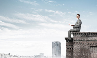 Man on roof edge reading book and cityscape at background