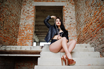 Brunette plus size sexy woman, wear at black leather jacket, lace panties, bra and high heels with chain at hands, siting on stairs at abadoned place. BDSM theme.