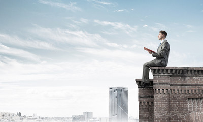 Man on roof edge reading book and cityscape at background