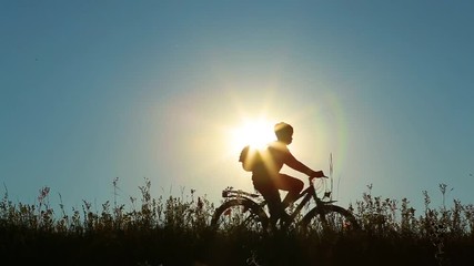 Little kid riding bike in summer meadow landscape at beautiful sunset sky background. Silhouette of anonymous caucasian small boy with backpack on back sitting on bicycle in sunset background.
