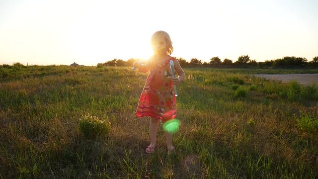 Little girl blowing soap bubbles against the sunset background. A child in a dress, a summer shot in a slow-motion picture.