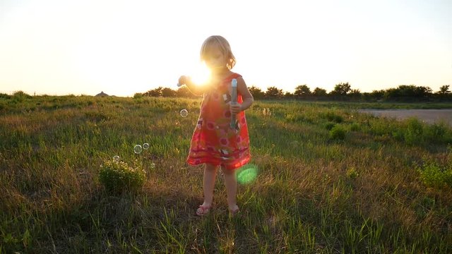 Little girl blowing soap bubbles against the sunset background. A child in a dress, a summer shot in a slow-motion picture.