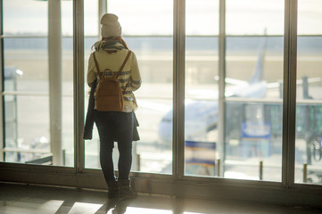 A woman with a backpack behind her is standing by the big window at the airport.