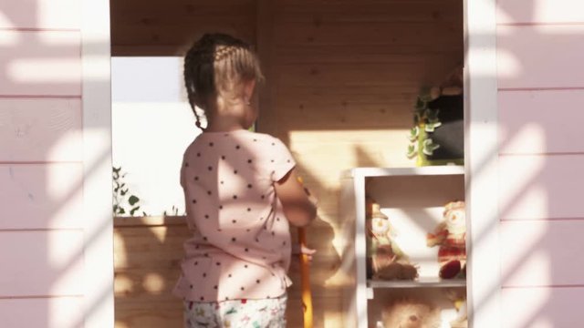 Backyard playground kids play house. Children playing inside wooden pink playhouse. Little girls pick up their toys and clean up the room of cubbyhouse after play.