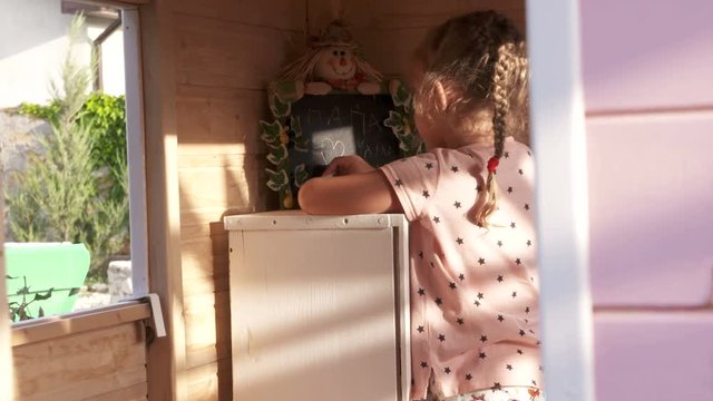 Little girl playing inside pink wooden play house on playground in the backyard. Child writing phrase in Russian on the blackboard I love Mom and Dad.