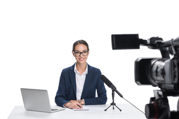 attractive female newscaster sitting at table with laptop, notepad and microphone looking at camera, isolated on white