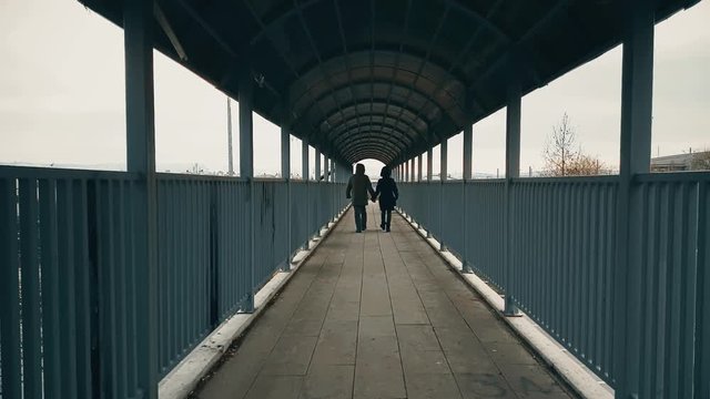 Young Couple With Coat Walking Hand-to-hand Across The Footbridge Away From The Camera Over Highway