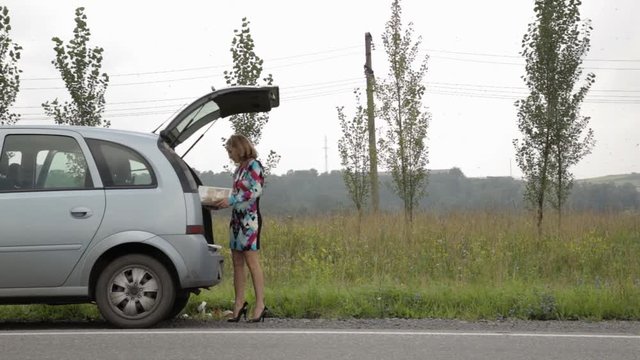 blonde girl stending near her broken car with open trunk on a country road and read the instruction