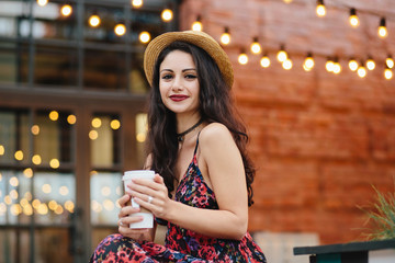 Outdoor shot of pretty female with dark long hair, having pure healthy skin, wearing straw hat and dress, drinking hot tea while sitting at terrace of cozy cafeteria. Beautiful woman resting at cafe