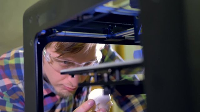 A man holds a tiny doll head sample and smiles at a working 3d-printer. 4K.