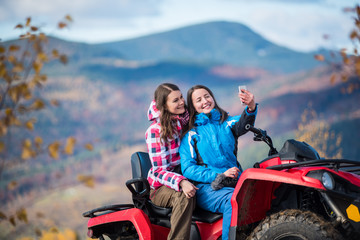 Fototapeta na wymiar Close-up two brunettes women in winter jackets on red quad bike smiling and makes selfie on the phone with blurred background nature