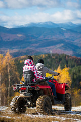 Rear view of couple in winter clothing driving a quad bike atv at the hill. Beautiful mountains and forests on the background