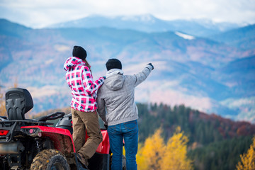Rear view of young pair near atv. Man is showing something in distance to her girlfriend. Blurred autumn landscape mighty mountains and forests on background © anatoliy_gleb