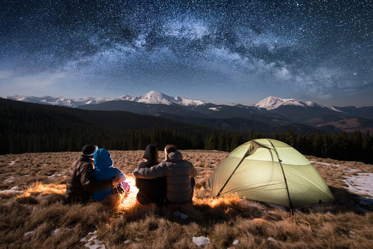 Silhouette of four people - two romantic couples are sitting together beside camp and tent under beautiful night sky full of stars and milky way. On the background snow-covered mountains. Rear view