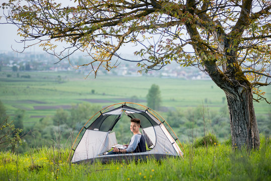 Young woman smiling to the camera while using her laptop outdoors sitting in a tent on top of a hill under tree while camping travelling journey weekend trip nature positivity studying