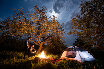 Romantic couple tourists standing at a campfire near tent under trees and night sky with the moon. Night camping