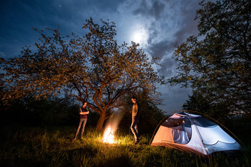 Romantic couple tourists standing at a campfire near tent under trees and night sky with the moon. Night camping