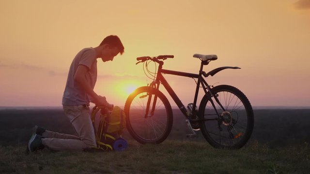 A young man puts things in a backpack. Sits by the bike at sunset