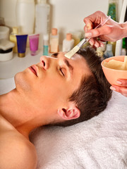 Obraz na płótnie Canvas Mud facial mask of man in spa salon. Massage with clay full face. Guy on with therapy room. Man lying wooden spa bed. Rejuvenating rest in the elite spa salon. Anti-aging cosmetic mask.