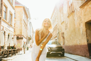 Young beautiful cheerful tanned woman in white dress walking on old street at tropical town. Pretty girl looking at you and smiling