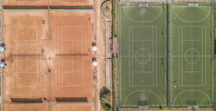 Vista aerea perpendicolare di due campi di calcio a 5 e un campo da tennis in terra rossa. A bordo campo panchine e ombrelli. I campi sono vuoti perch&egrave; &egrave; estate e fa caldo.