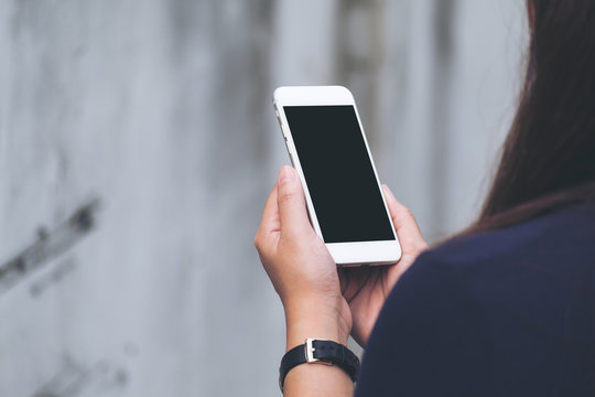 Mockup image of a woman holding and using white smart phone with blank black screen in outdoor and old white concrete wall background