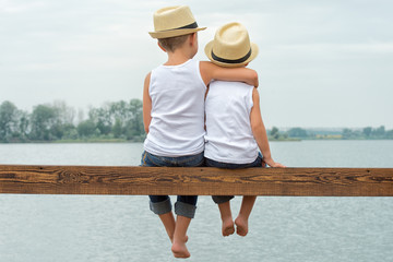 Two brothers in a straw hats sitting on the pier and looking at the lake.Summer vacation 