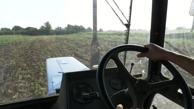 Farmer in hat, sits inside in the tractor cabin, rides on the field with plow.