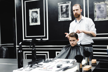 Young handsome barber making haircut of cute boy in barbershop