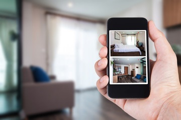 Hand holding smartphone view looking in house on blurred house background with Home security CCTV system concept.