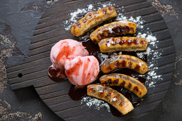 Top view of a black wooden serving board with fried mini bananas, ice cream, sauce and powdered sugar