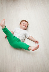 happy baby boy in white t-shirt and green pants lying on her back on a wooden floor