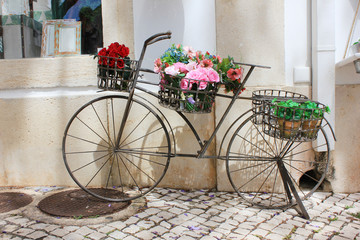 Fototapeta na wymiar Outdoor creative decoration of old fashioned vintage metal bicycle with colorful flowers, plant pot on cobblestone pavement. European cozy small street lifestyle every day scene in shopping district.