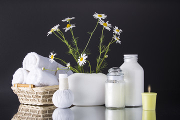 Spa. Chamomile in a vase, three white towels rolled into a roll lie in a basket. Two cans of cream. Candle and herbal balls. The background is black.