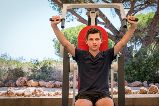 Young caucasian male exercising upper body on gym machine outdoors surrounded by trees.