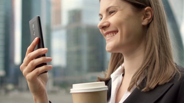 Young businesswoman in suit using smart phone in city downtown, professional female having video chat conference recieving good news talking live-streaming. Business district skyscrapers in background