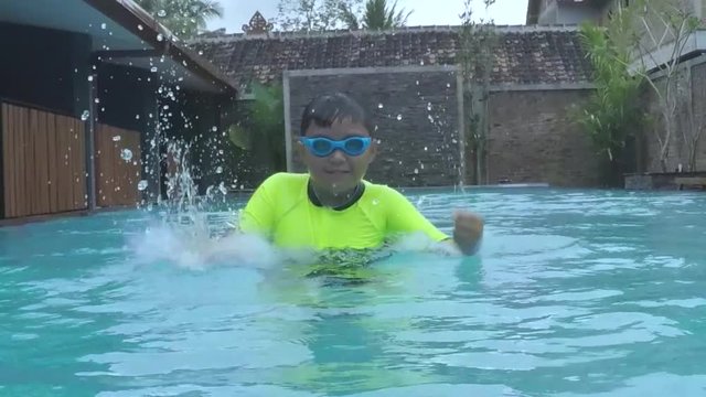 Slow motion footage of a cheerful little boy splashing water in the swimming pool while wearing swimsuit and goggles