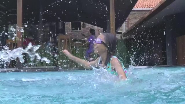 Slow motion footage of a cheerful toothless girl splashing water in the swimming pool while wearing a swimsuit and goggles