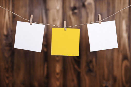 Close up of two white and one yellow note papers hung by wooden clothes pegs on a brown wooden background