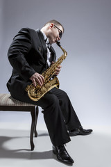 Naklejka premium Music Concepts. Portrait of Mature Relaxed and Thoughful Caucasian Saxophone Player in Sunglasses Playing the Saxophone While Sitting on Chair in Studio Environment.