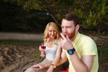 Obraz premium Young man and girl sitting and hugging in the woods and drink wine. Young beautiful couple smiling and drinking wine on picnic in summer Park on green grass near forest