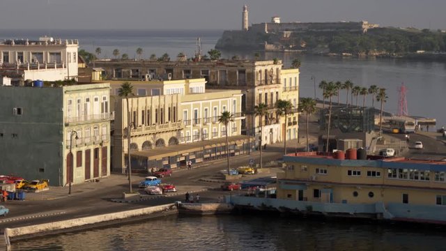A high angle long establishing shot of the restaurants and bars along the shoreline of Havana, Cuba harbor. The lighthouse at Faro del Castillo del Morro is in the distance. Shot in 5K.  	