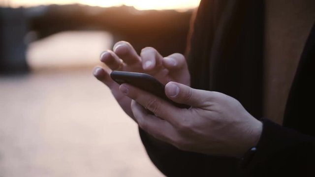 Close-up view of male hands holding the smartphone and typing on touchscreen, standing on embankment on sunset.