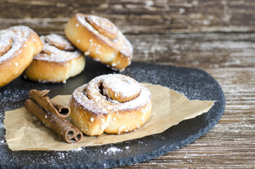 Delicious sweet cinnamon rolls with powdered sugar and cinnamon stick in baking paper on wooden table. Sweet food backround..